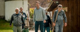 Schloss Tannegg Eine Gruppe von fünf Männer laufen einen schmalen Kiesweg hinunter und tragen ihr Werkzeug für die Gartenarbeit mit sich.