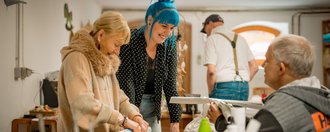 Schloss Tannegg Eine Mitarbeitende steht mit einer Gruppe Bewohnenden in der Kreativwerkstatt des Hauses, diese Rollen mit viel Freude Zeitungspapier zusammen, für ein bevorstehendes Bastelprojekt.